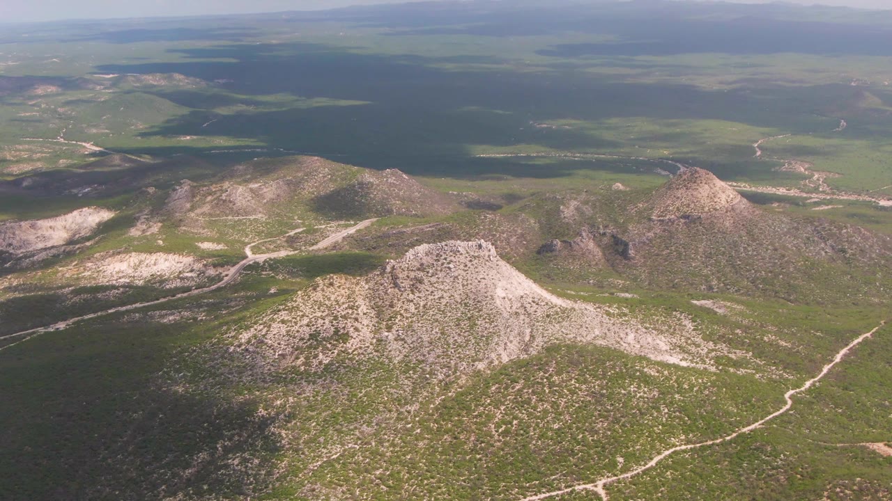 Opening aerial frame over Buzzards Beach East Cape BCS Mexico terrain, with a pale rocky hill in the foreground, green plains beyond, distant ridges, cloud shadow, and no visible people.