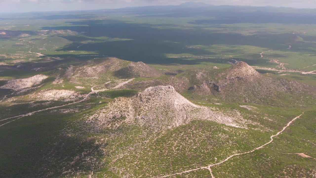 Aerial frame over the East Cape BCS Mexico landscape, showing a rounded pale hill, darker cloud-shadowed ground to the left, scattered rocky peaks, and no visible people.