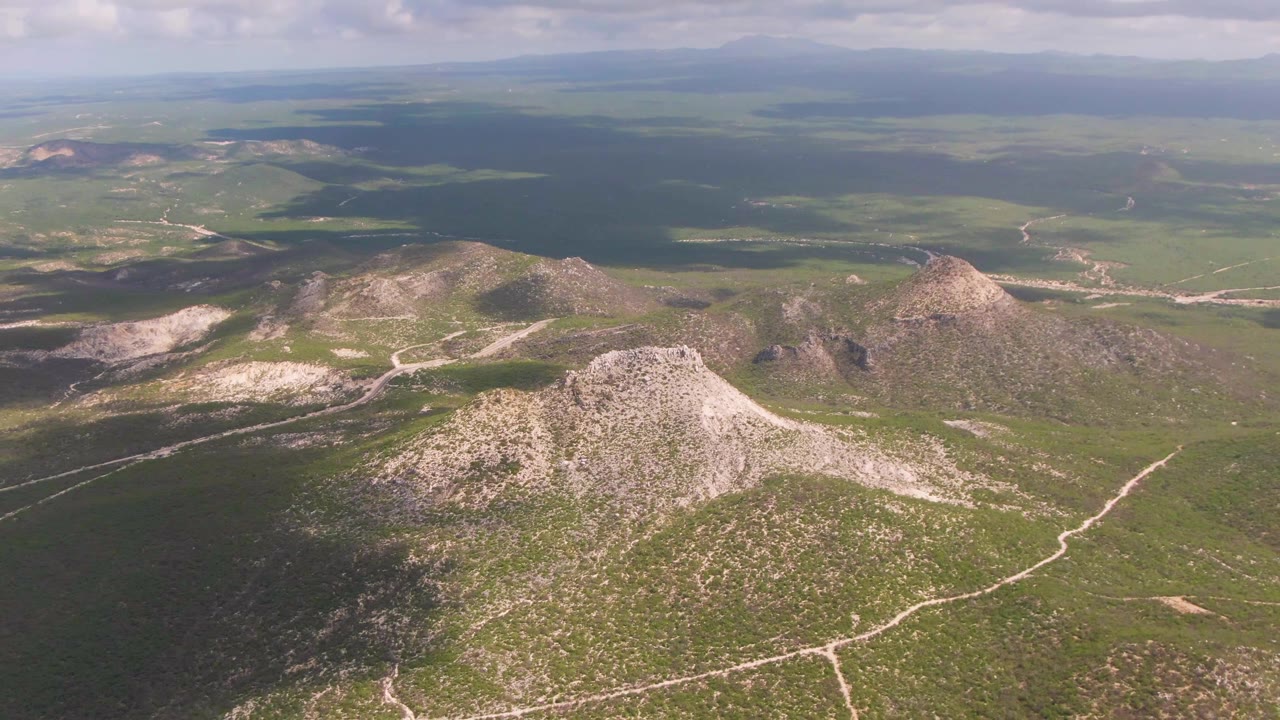 High aerial view from Buzzards Beach East Cape BCS Mexico, with a central rocky hill, intersecting dirt roads on green terrain, distant flatlands, and no visible people.
