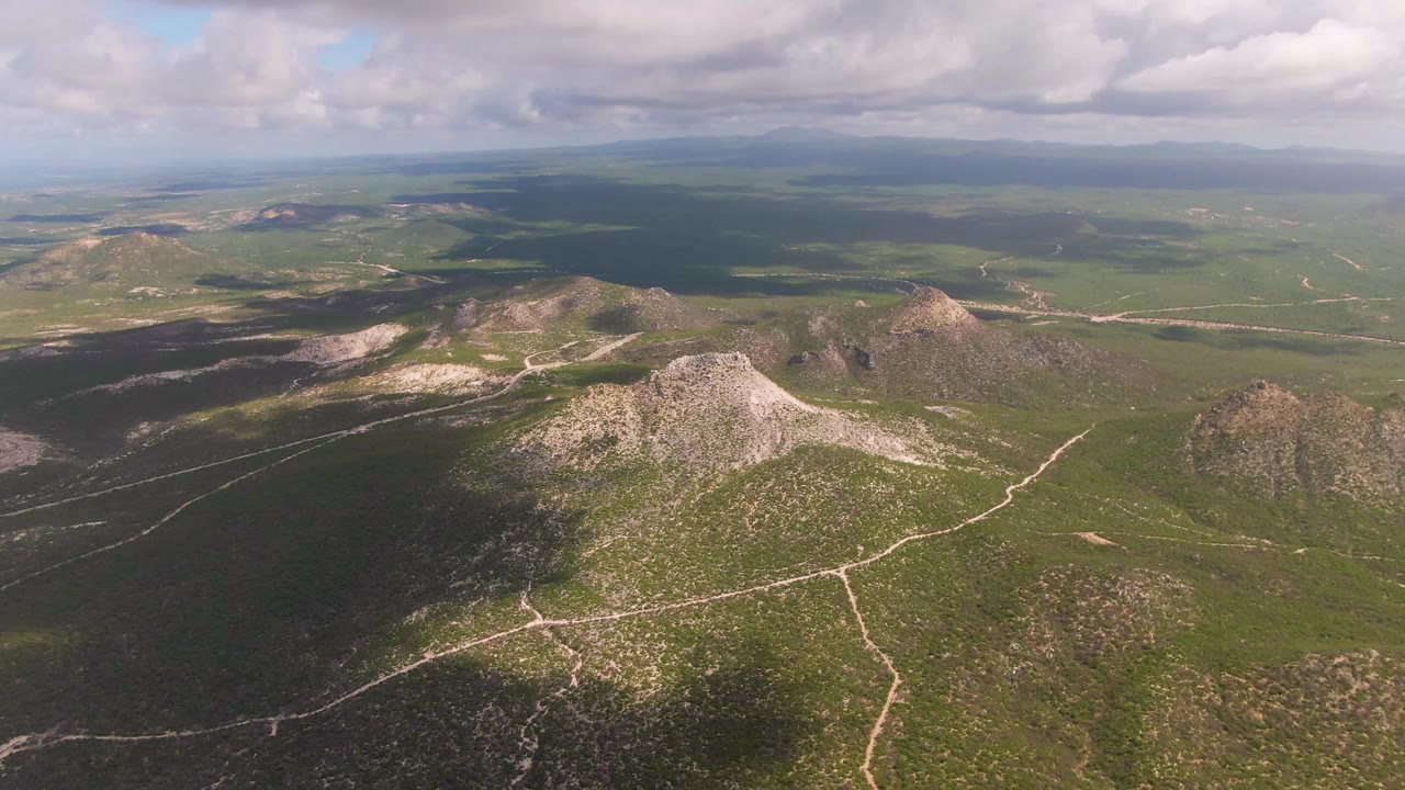 Aerial frame from the Buzzards Beach East Cape BCS Mexico collection showing rocky midground hills, branching dirt tracks, green scrub terrain, heavy cloud shade to the left, and no visible people.
