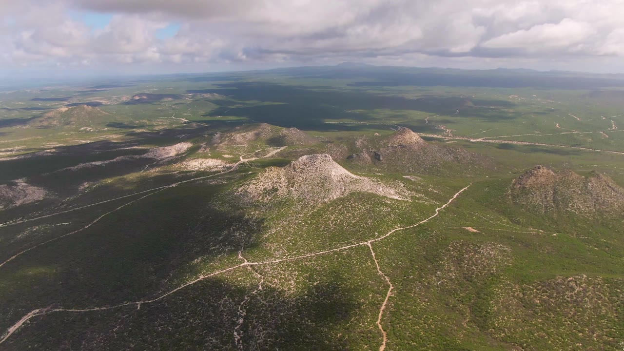 Wide drone view over Buzzards Beach East Cape BCS Mexico backcountry, with a central rocky ridge, green lowlands, pale tracks, layered distant hills, broken clouds, and no visible people.