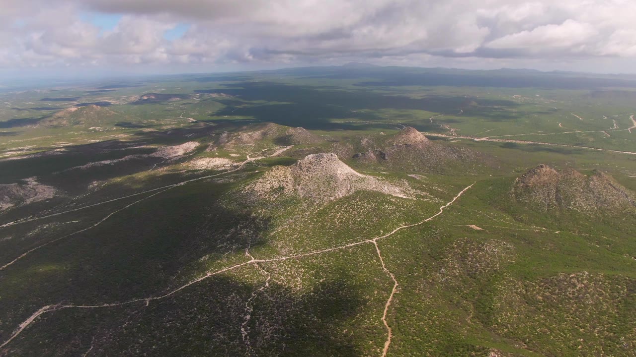 Aerial landscape of green valleys, rocky hills, dirt roads, and a broad cloud bank.