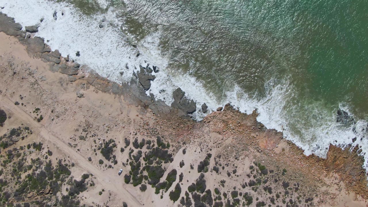 Overhead view of Buzzards Beach East Cape BCS Mexico with green water, white surf, dark rocks, tan sand, scrub vegetation, and no visible people.