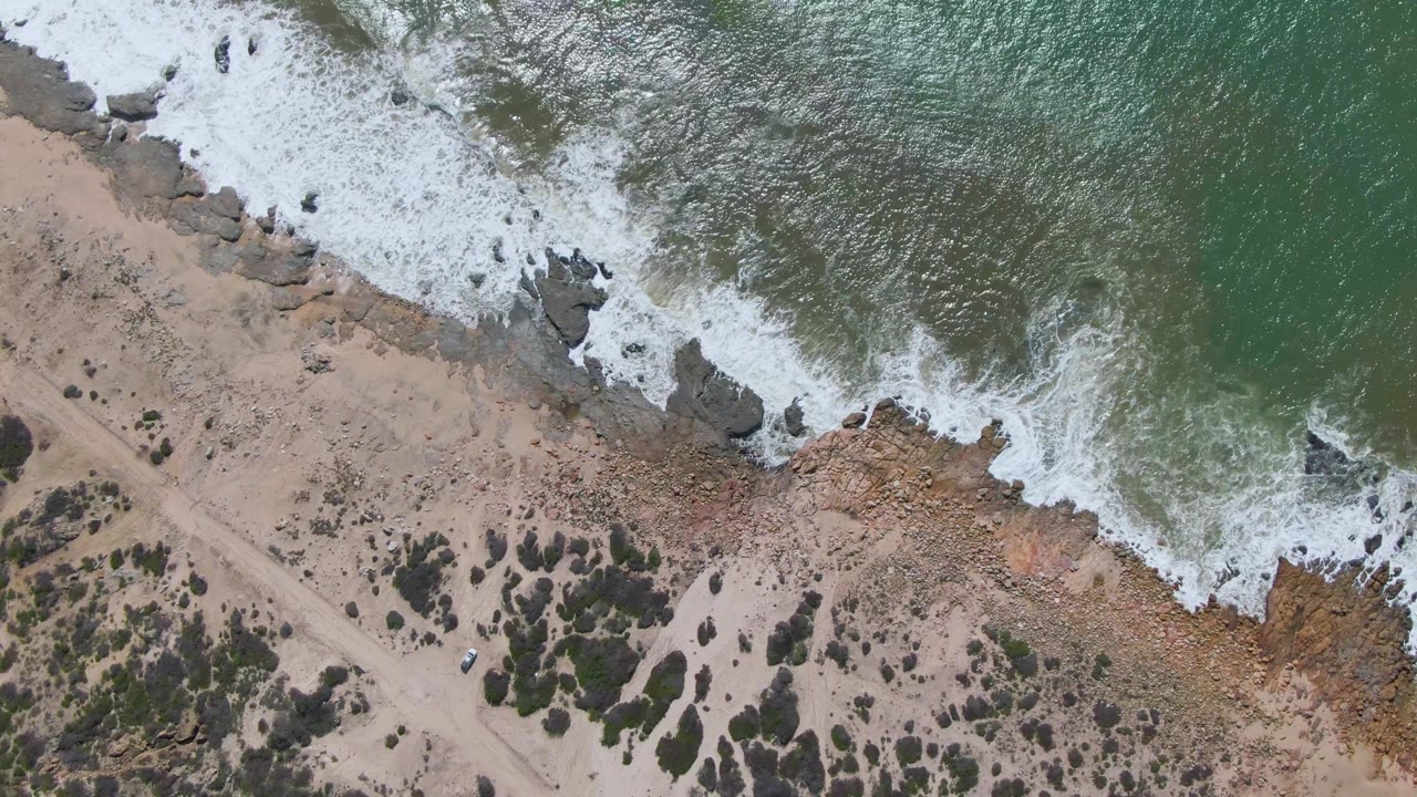Aerial frame over Buzzards Beach East Cape BCS Mexico showing foam spreading over rocks, sandy beach scrub, a dirt path, a small vehicle, and no visible people.