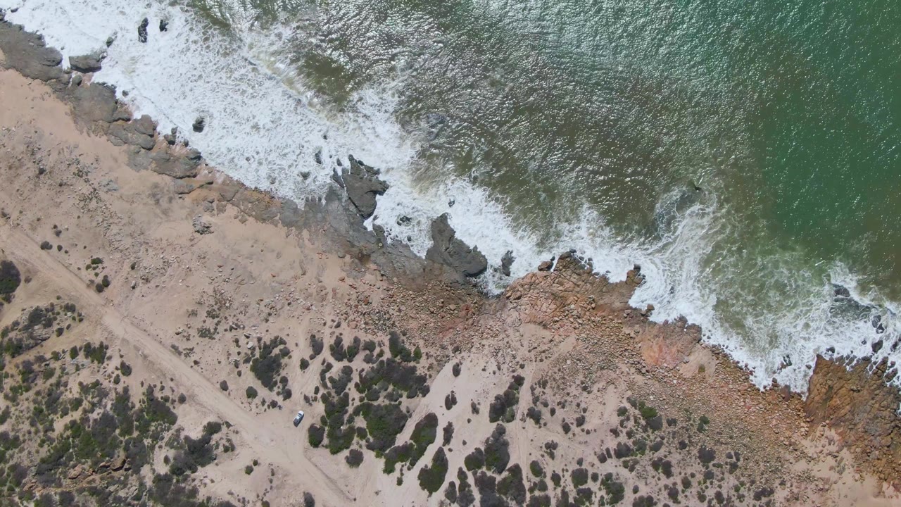 Top-down view of Buzzards Beach East Cape BCS Mexico where white water presses farther along the rocky edge beside tan sand, low vegetation, a faint track, and no visible people.