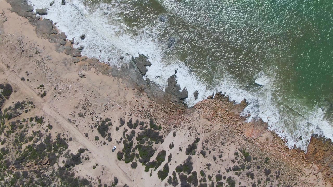 Overhead view of Buzzards Beach East Cape BCS Mexico with white surf breaking over dark rocks, green-blue water, tan sand, a dirt path, sparse scrub, and no visible people.