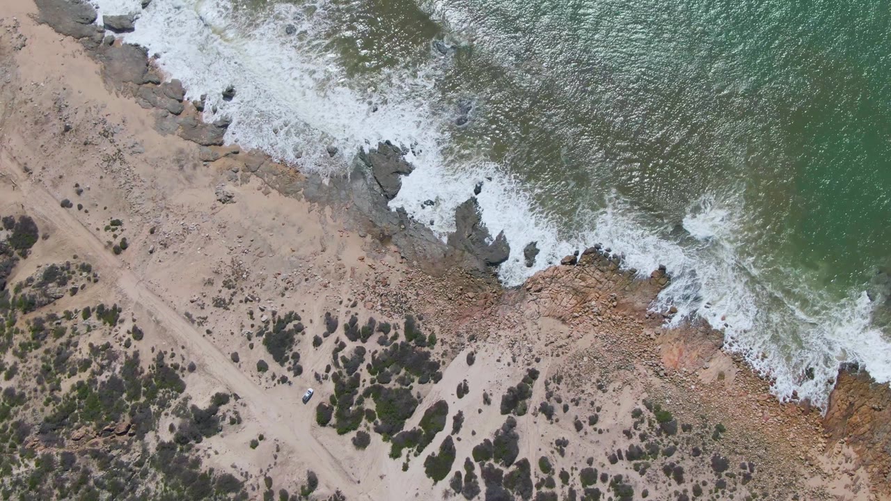 Aerial frame over Buzzards Beach East Cape BCS Mexico as whitewater spreads along a jagged rocky edge beside sand, scrub patches, a dirt track, and no visible people.