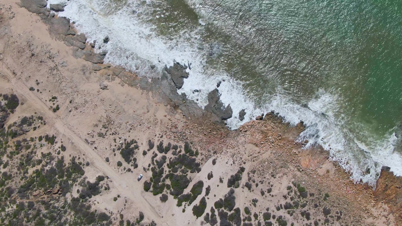 Buzzards Beach East Cape BCS Mexico seen from above, with foamy waves curling into shoreline rocks, tan beach, scattered dark shrubs, a pale dirt path, and no visible people.
