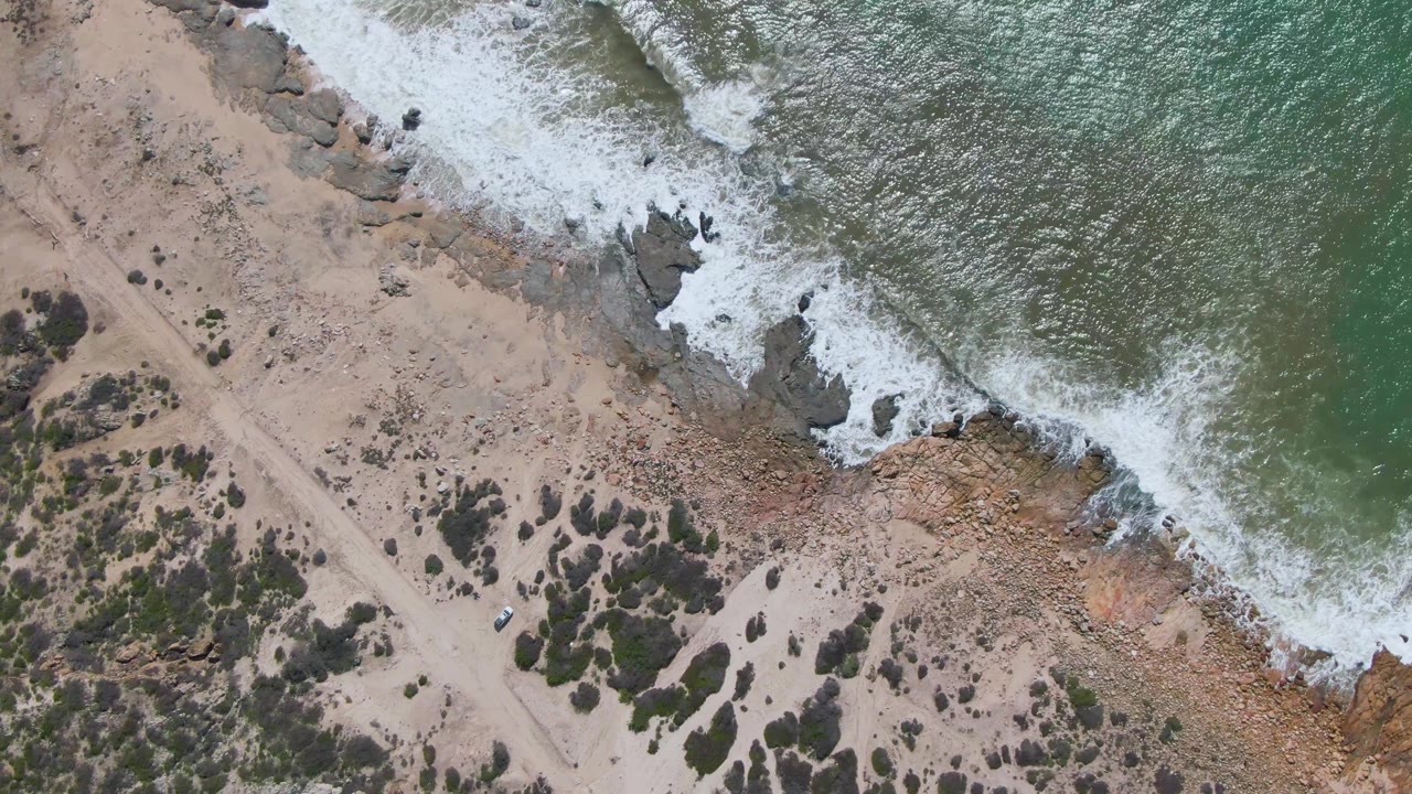 Aerial view of waves, rocky shore, sandy road, scrub, and a small vehicle.