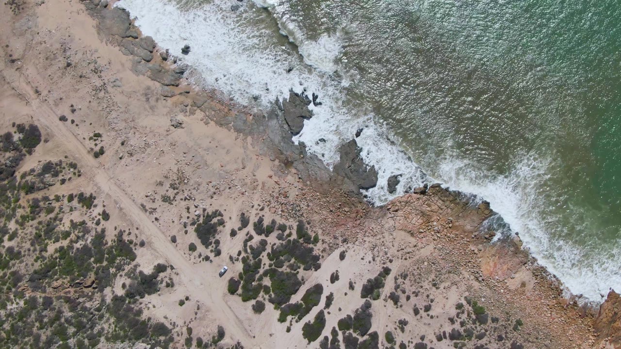 Aerial frame at Buzzards Beach East Cape BCS Mexico with whitewater spreading along the rock edge, the pale dirt road running through sand, and the small vehicle slightly farther along, no visible people.
