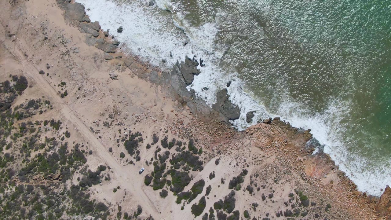 Overhead view at Buzzards Beach East Cape BCS Mexico showing the green ocean occupying the right side, foam crossing the rocks, and the sandy access road with scattered shrubs on the left, no visible people.