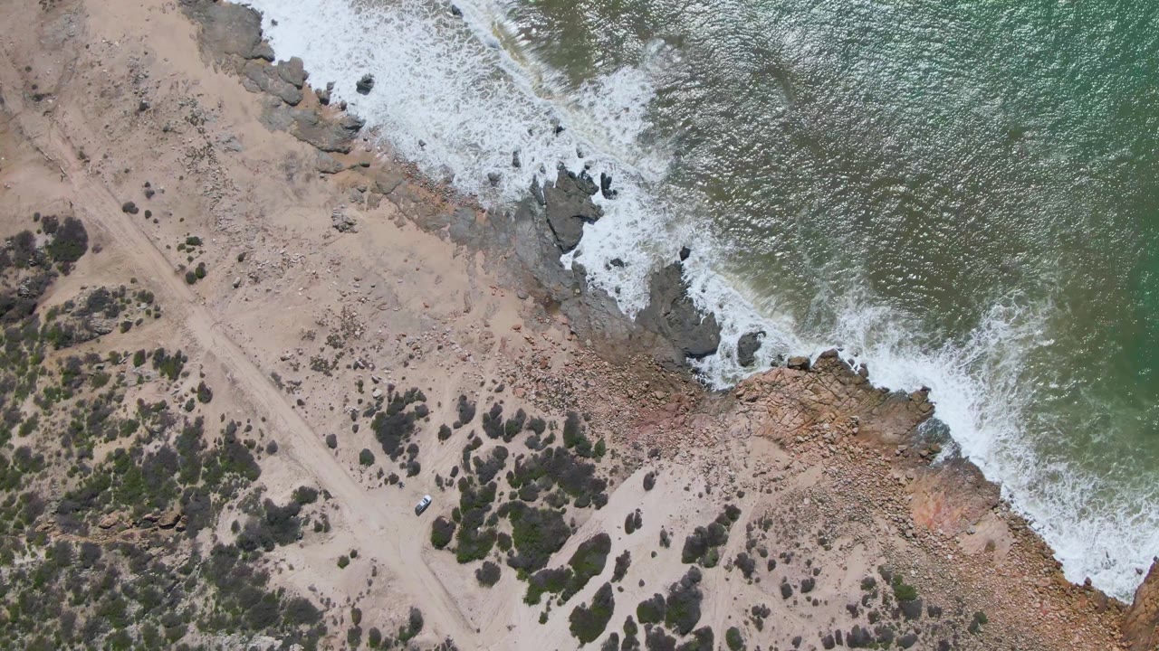Aerial frame at Buzzards Beach East Cape BCS Mexico where the surf line sits higher in the composition, the road cuts diagonally through the sand, and brush clusters fill the lower left, no visible people.