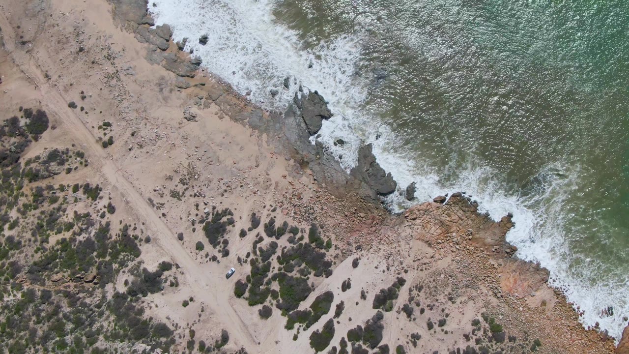 At Buzzards Beach East Cape in Mexico, a top-down aerial frame shows green water foaming over rocks beside tan sand, a dirt road, scrub vegetation, a small vehicle, and no visible people.