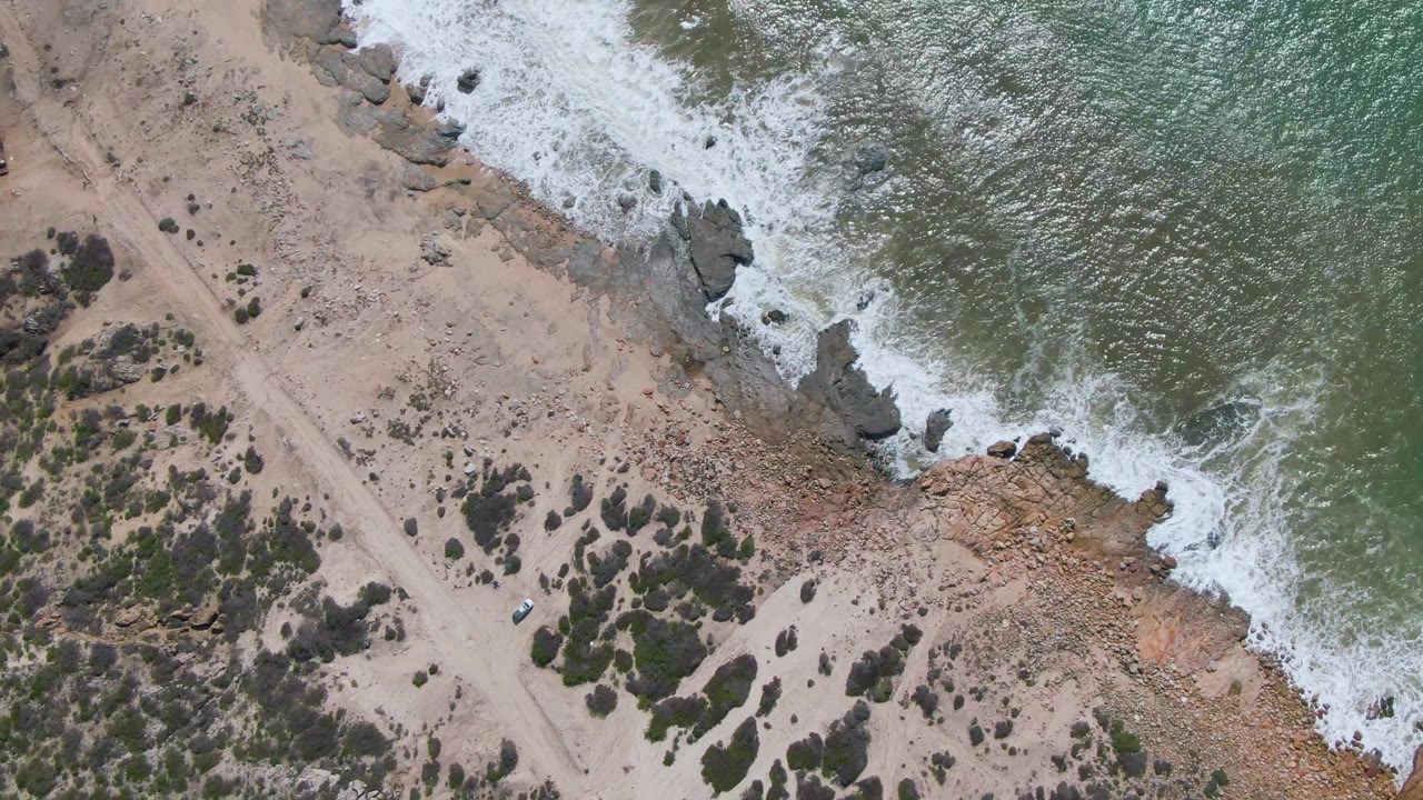 At 0:01, the Buzzards Beach East Cape shoreline fills the frame with white surf pushing farther along the rocks, while the dirt road and small vehicle remain inland with no visible people.
