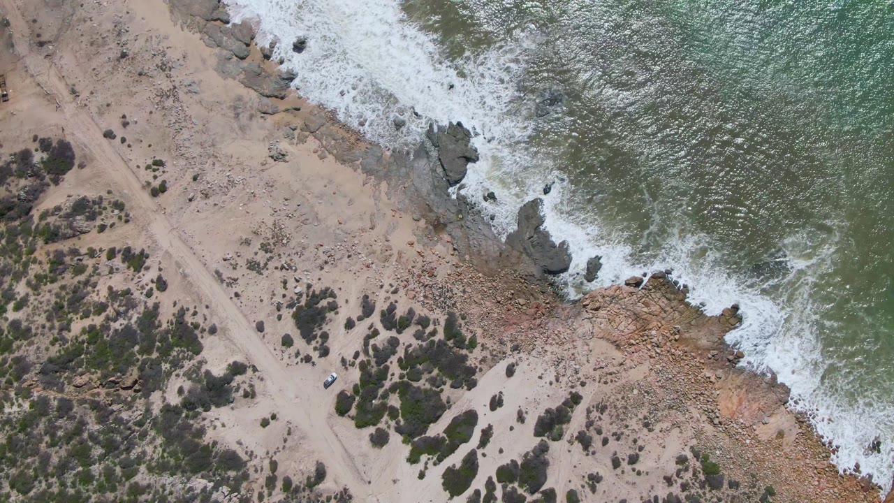 At 0:02, the aerial view over Buzzards Beach East Cape shows rippled green water, fresh foam on the rocky edge, a pale dirt road, scattered shrubs, a small vehicle, and no visible people.