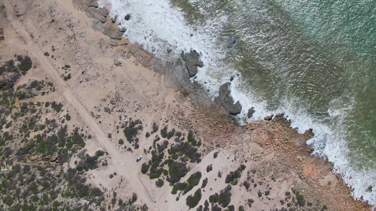 Aerial frame of a sandy coastal road below surf and rocky shoreline.