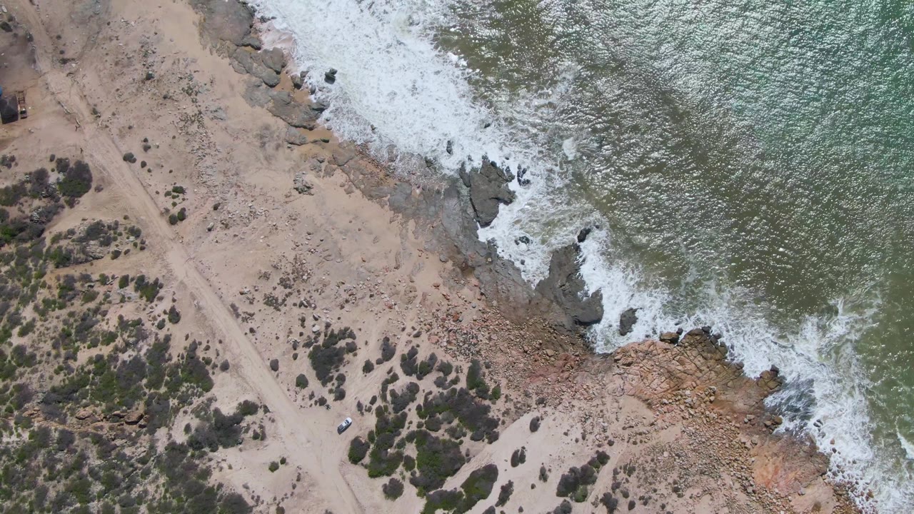 At Buzzards Beach in Mexico, an overhead view shows white surf meeting rocky shoreline beside a sandy path with scrub bushes, a small blue-roof structure near the top left, a white vehicle near the lower left, and no visible people.