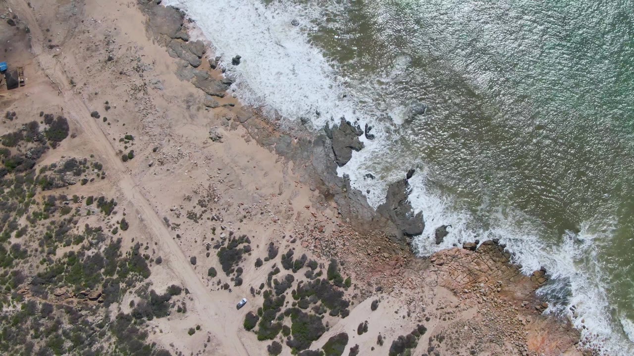 At Buzzards Beach in Mexico, the aerial frame shows the white vehicle farther along the sandy track, with green-gray water and white foam pressing against dark shoreline rocks and no visible people.