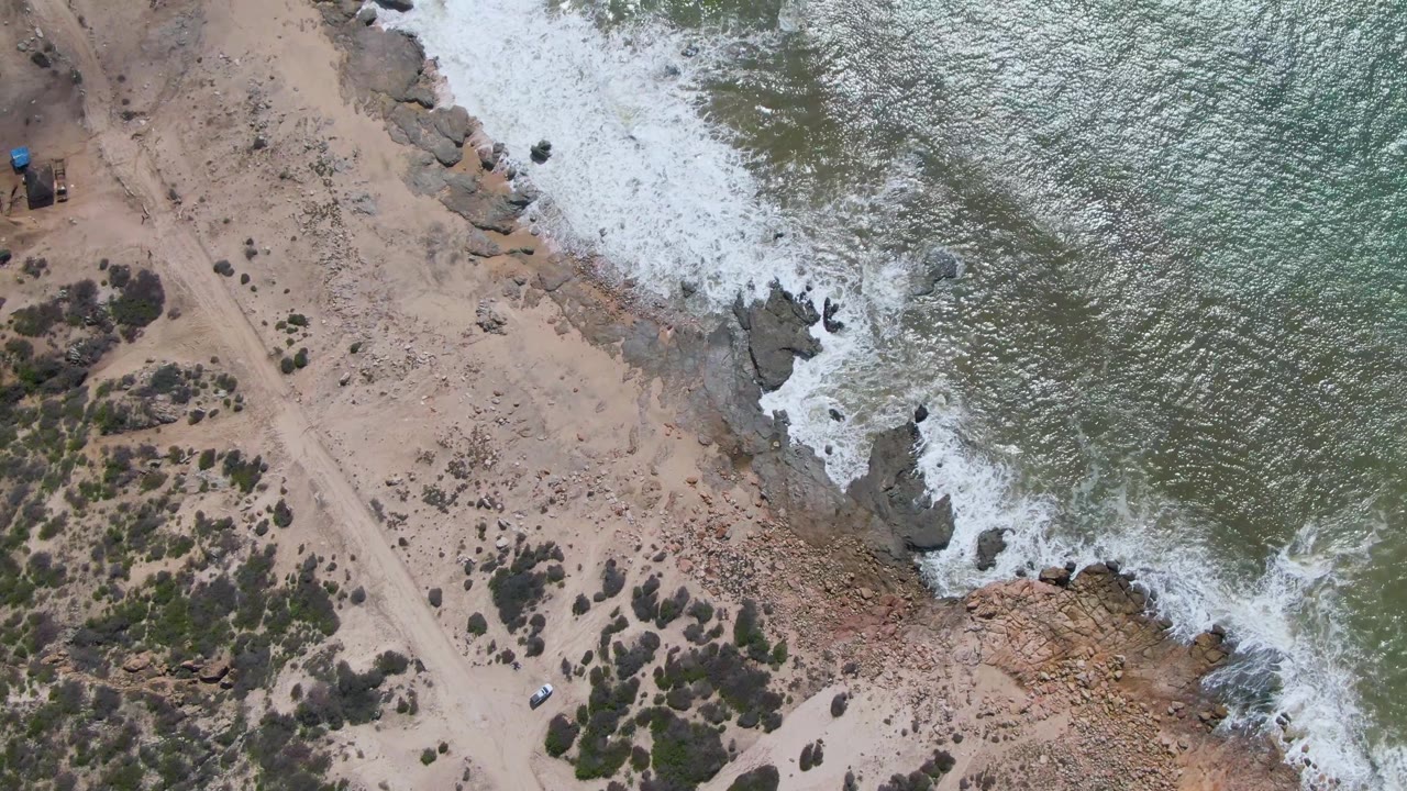 At Buzzards Beach in Mexico, the top-down frame places the sandy road through the center with scattered shrubs, the blue-roof structure near the upper left, the white vehicle near the road, and surf curling over rocks to the right with no visible people.