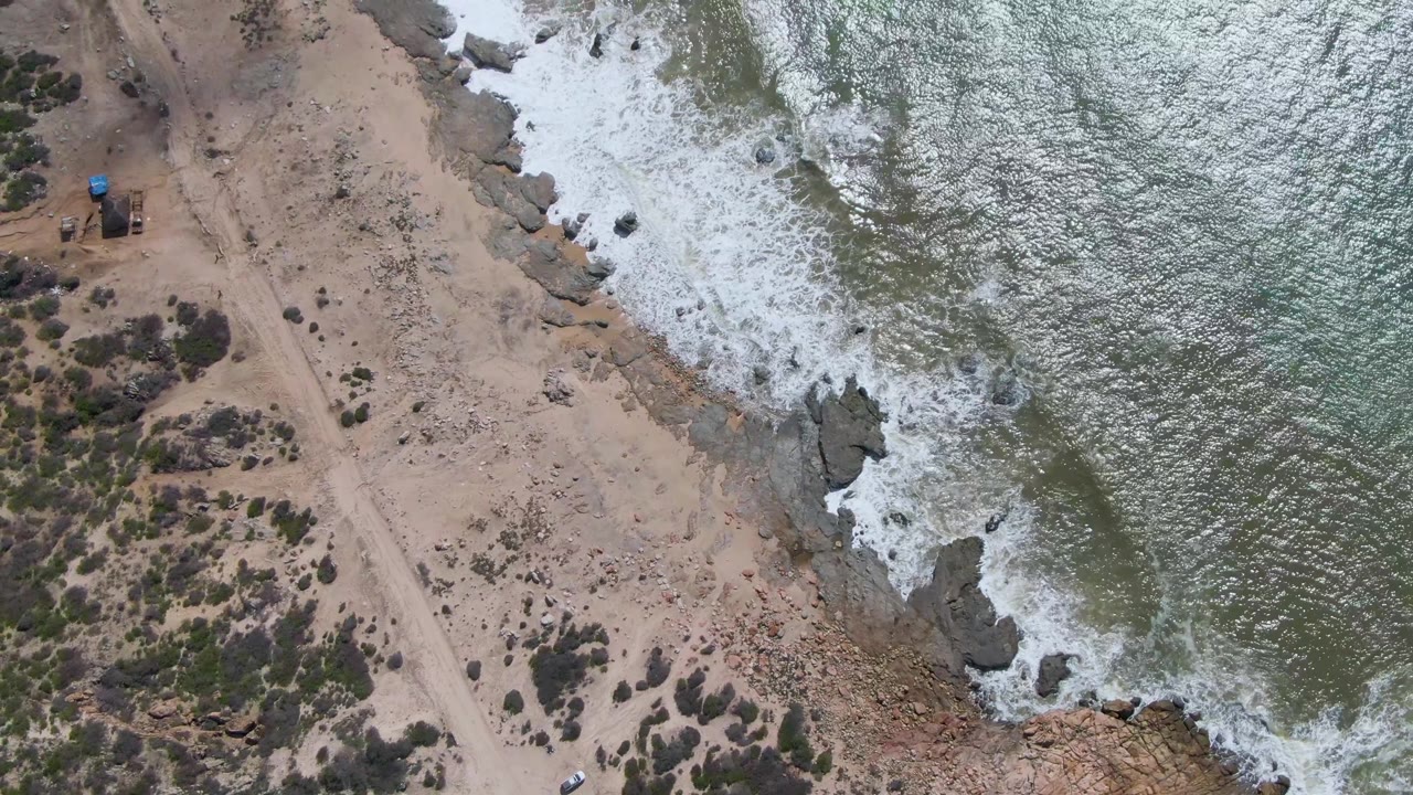 Overhead coastal dirt road beside rocky surf and scrubland.