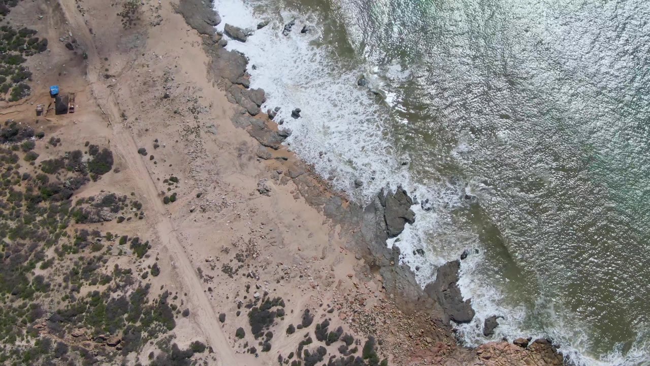 Aerial frame at Buzzards Beach East Cape BCS Mexico with white surf spreading along the rocks beside an unpaved road and scrub-covered ground, with no visible people.