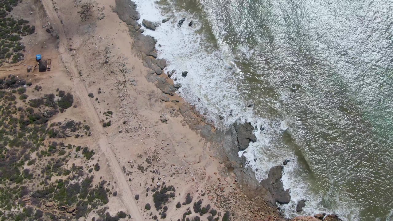 High overhead view at Buzzards Beach East Cape BCS Mexico showing foamy ocean water against shoreline rocks, a parallel dirt road, dry terrain, and no visible people.
