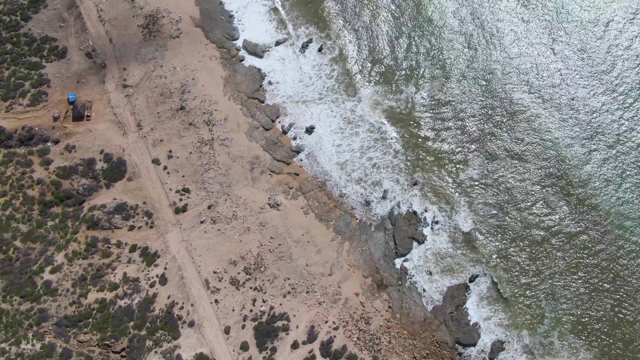 Buzzards Beach East Cape BCS Mexico appears from above with a dirt road crossing dry scrubland while whitewater curls around shoreline rocks and no visible people are present.