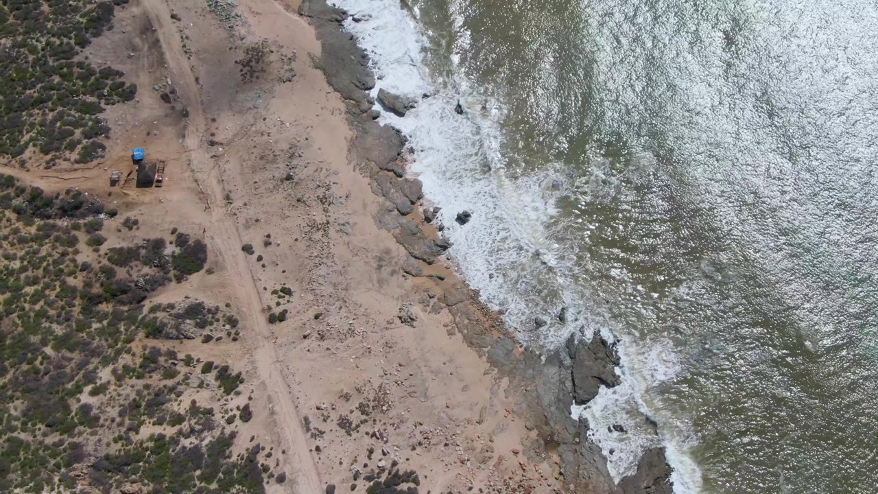 Aerial frame over Buzzards Beach East Cape BCS Mexico with a sandy dirt road on the left, rocky shoreline in the center, green-gray surf on the right, and no visible people.