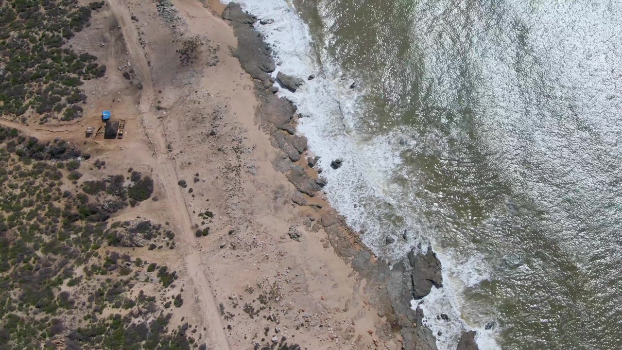 High drone view at Buzzards Beach East Cape BCS Mexico showing the dirt road and scrubland inland while white foam spreads around rocks along the waterline, with no visible people.