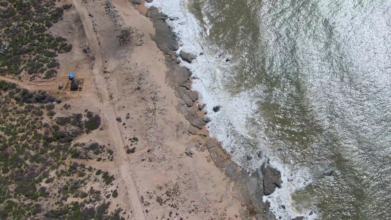 Drone frame over Buzzards Beach East Cape BCS Mexico with sun-glinted water on the right, a narrow rocky edge, pale sand, scrub patches, and no visible people.