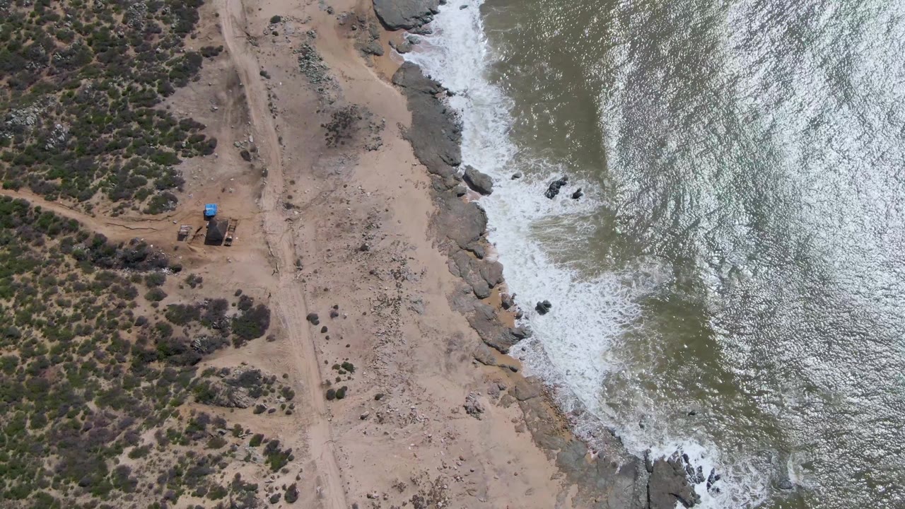 Overhead view of Buzzards Beach East Cape BCS Mexico with white surf breaking on rocks beside tan sand, sparse vegetation, a dirt path, a small blue-roofed structure, and no visible people.