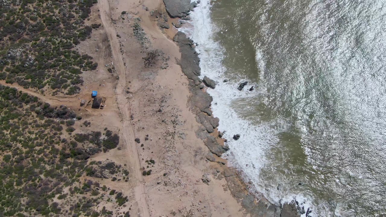 Aerial frame over Buzzards Beach East Cape BCS Mexico as a band of white foam spreads along the rock edge, with tan beach, scrub, a dirt track, and no visible people.