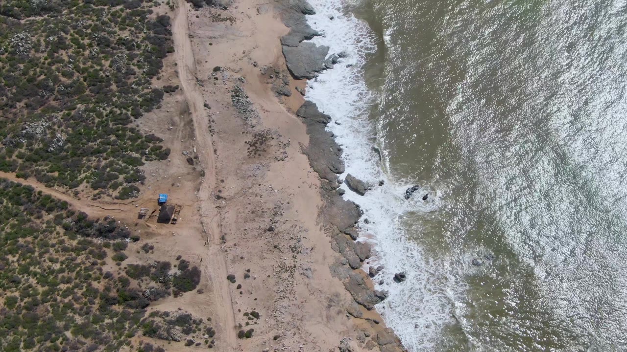 Aerial frame of Buzzards Beach East Cape BCS Mexico showing waves breaking higher along the rocky edge, with sand, scrub, a dirt path, and no visible people.