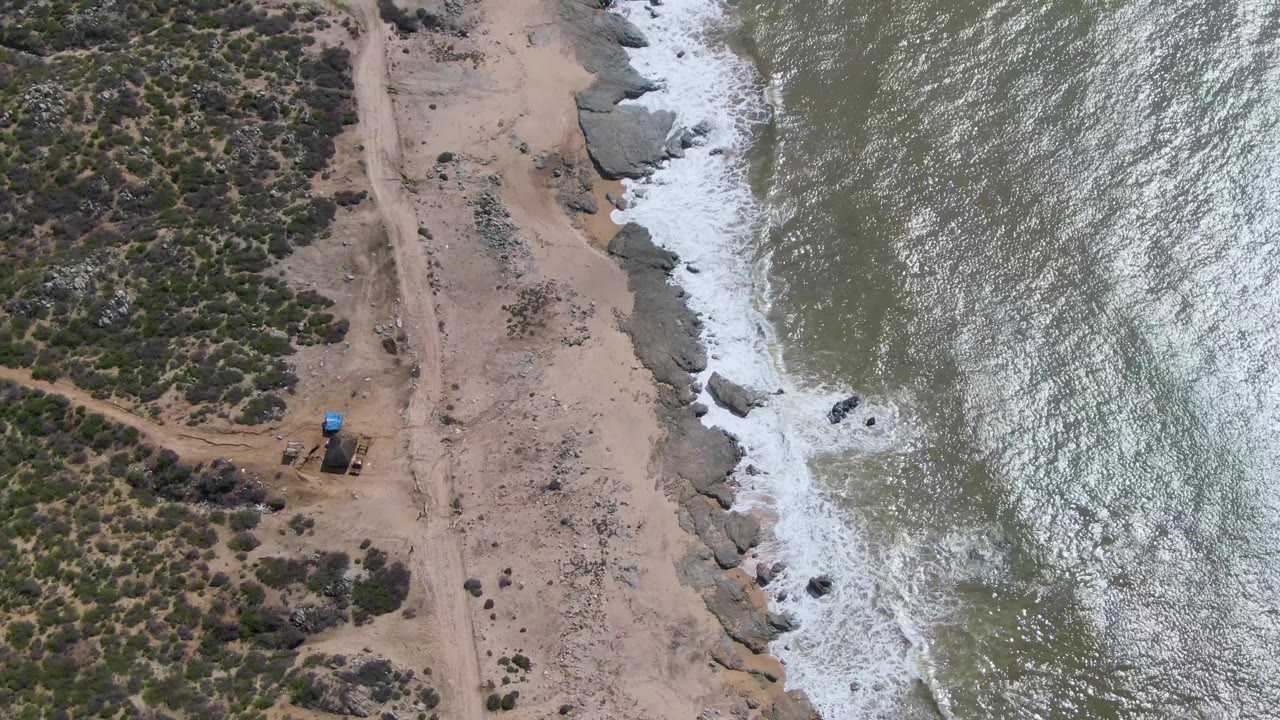 Drone view over Buzzards Beach East Cape BCS Mexico with bright reflected water, a foamy wave band, scattered rocks, dry vegetation, a dirt track, and no visible people.