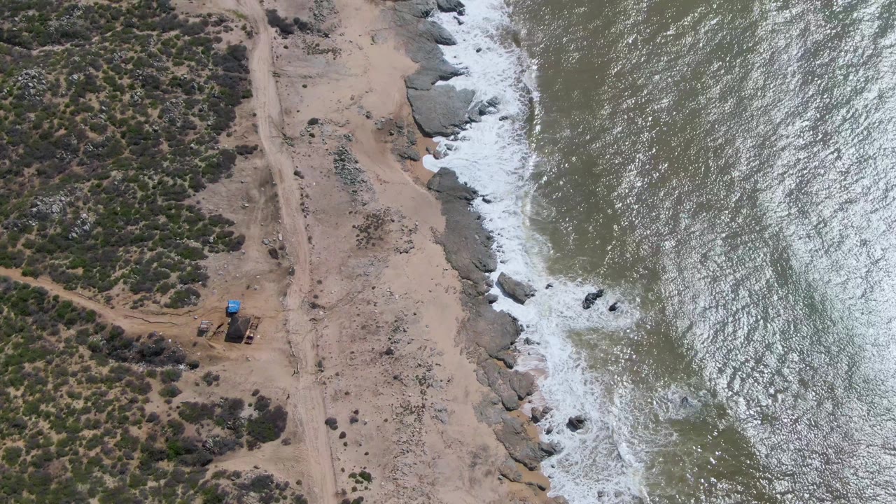 High-angle shoreline with ocean glare, foamy rocks, sand, and sparse vegetation.