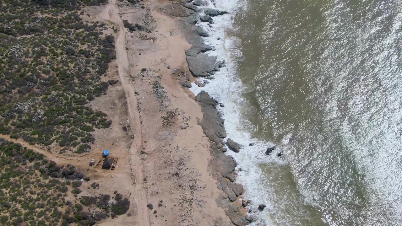 Opening aerial frame over Buzzards Beach in Mexico shows scrubland and a sandy dirt track beside rocks and breaking surf, with no visible people.