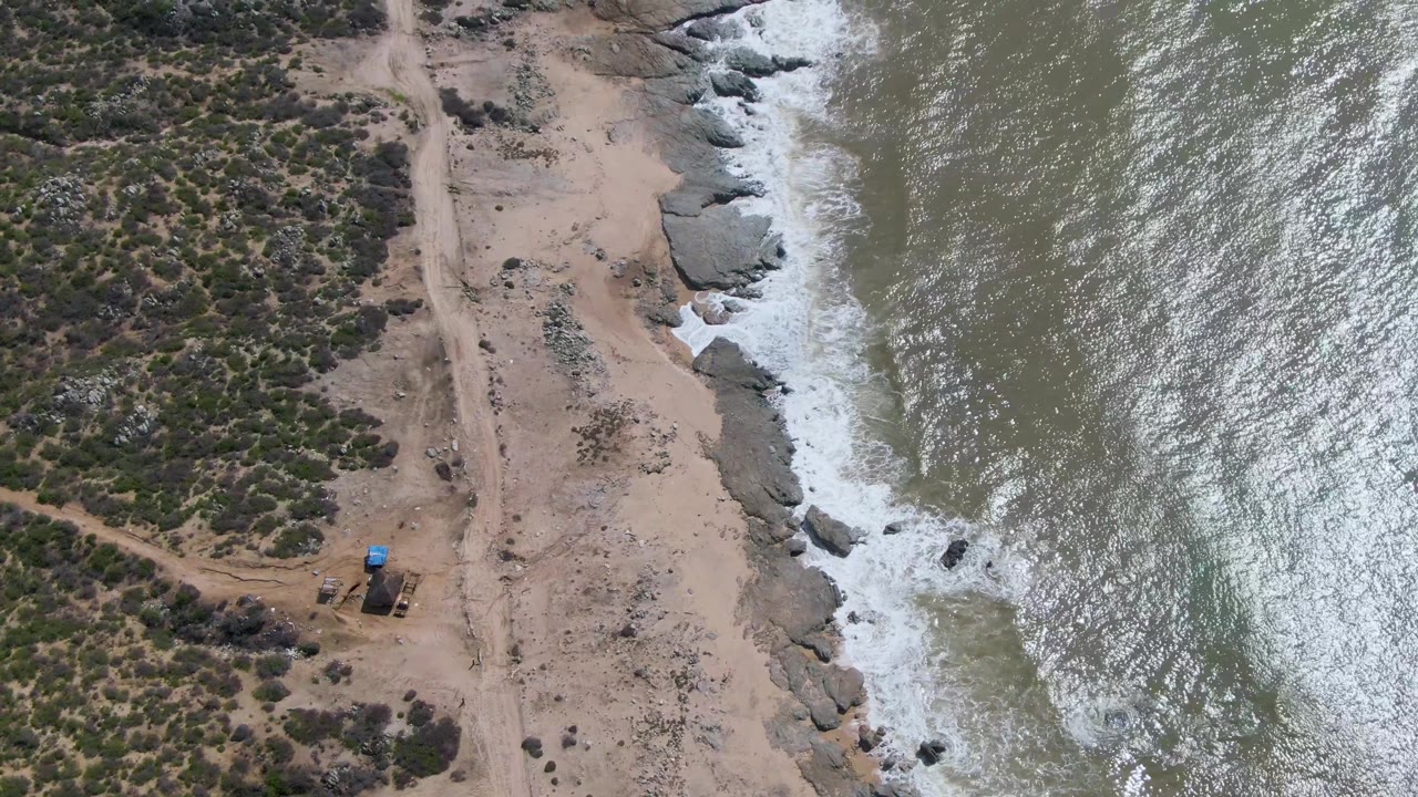At 0:01, the Buzzards Beach aerial view shows the dirt access path more centered beside pale sand, coastal rocks, foamy surf, and no visible people.