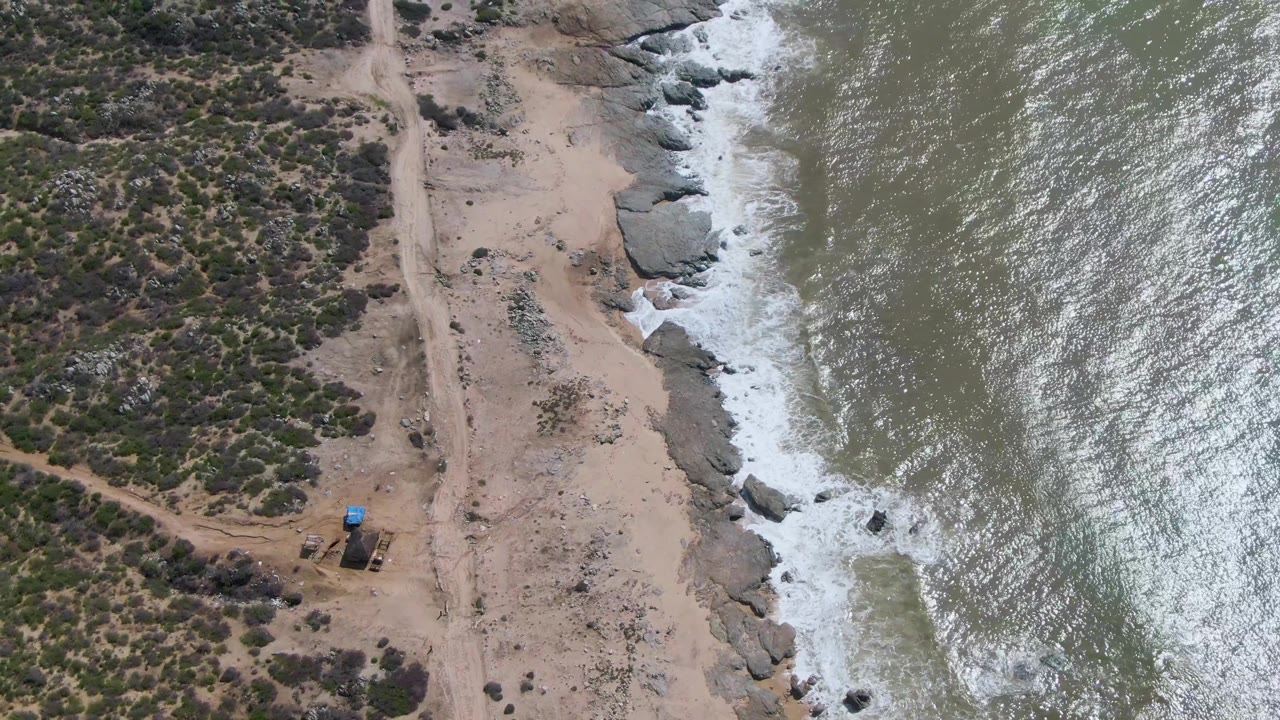 At 0:02, Buzzards Beach appears from above with scrub vegetation inland, a sandy track near shore, sunlit water on the right, and no visible people.