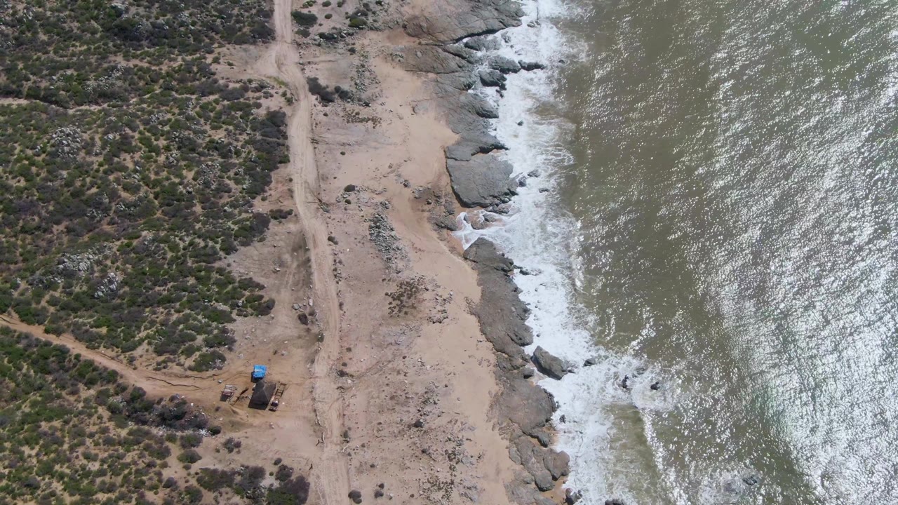 Aerial shoreline view with ocean waves, rocks, sandy ground, scrub, and a small vehicle.