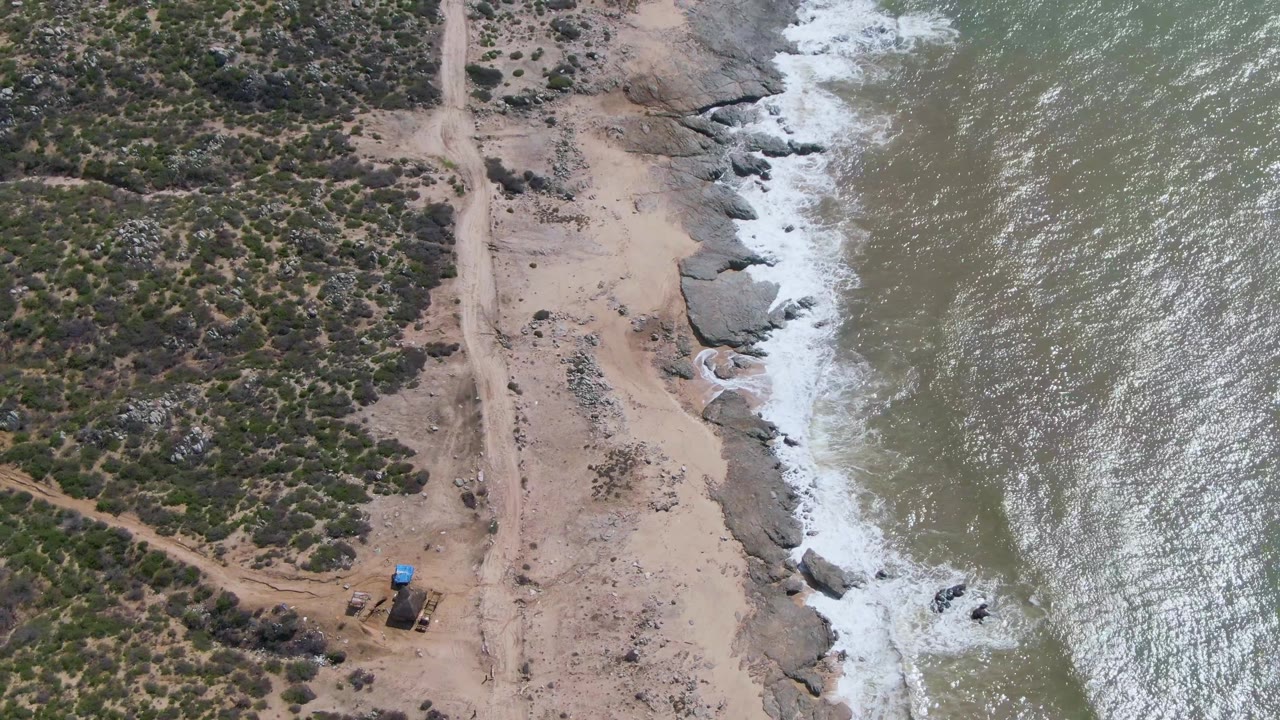 Aerial frame from Buzzards Beach East Cape BCS Mexico showing a dirt road between scrub-covered land and foamy waves on a rocky shore, with no visible people.