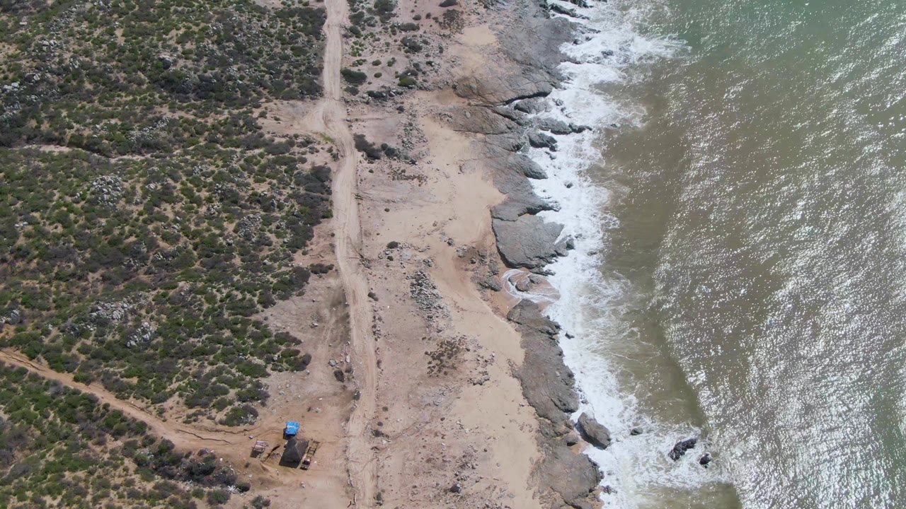 At 0:02, the aerial frame shows the East Cape shoreline with low green vegetation, a pale dirt road, exposed rocks, choppy green water, and no visible people.