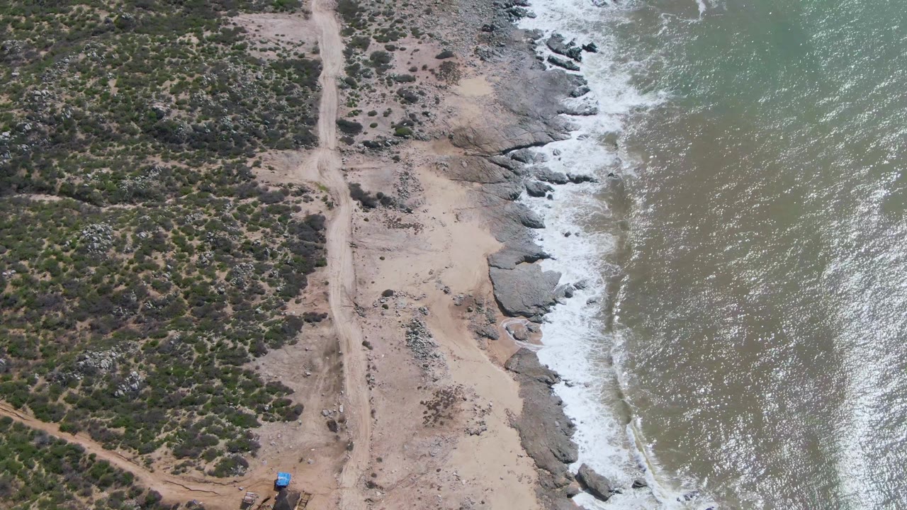 Aerial frame from Buzzards Beach East Cape BCS Mexico showing a sandy dirt road between scrubland and breaking surf, with a small blue vehicle near the lower left and no visible people.