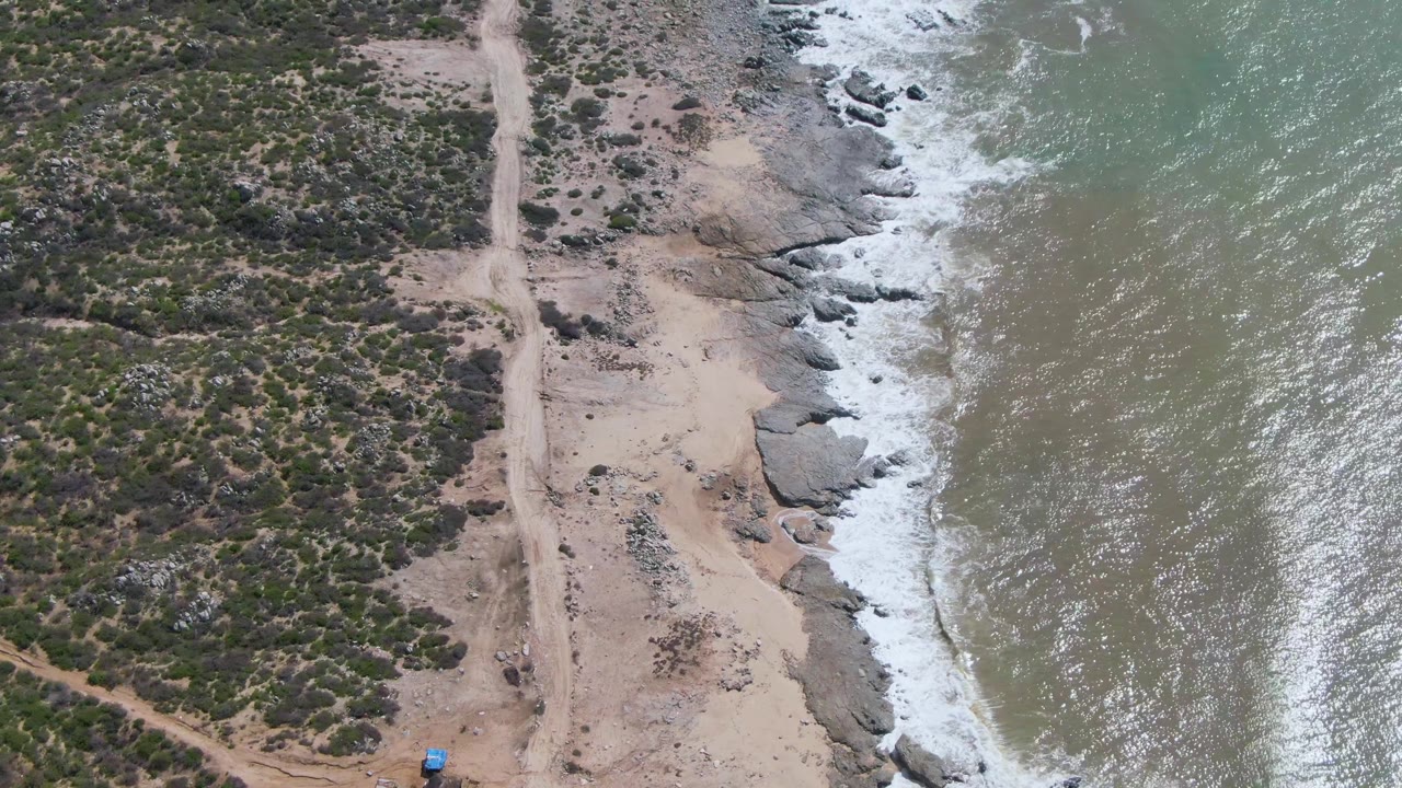 Aerial view at Buzzards Beach East Cape BCS Mexico with the dirt road centered, scrub vegetation to the left, foamy waves hitting rocks to the right, and no visible people.