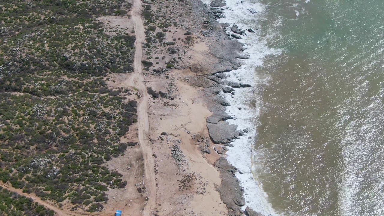 Drone frame over Buzzards Beach East Cape BCS Mexico showing scrub-covered land, a pale dirt track, exposed shoreline rocks, bright white wave foam, and no visible people.