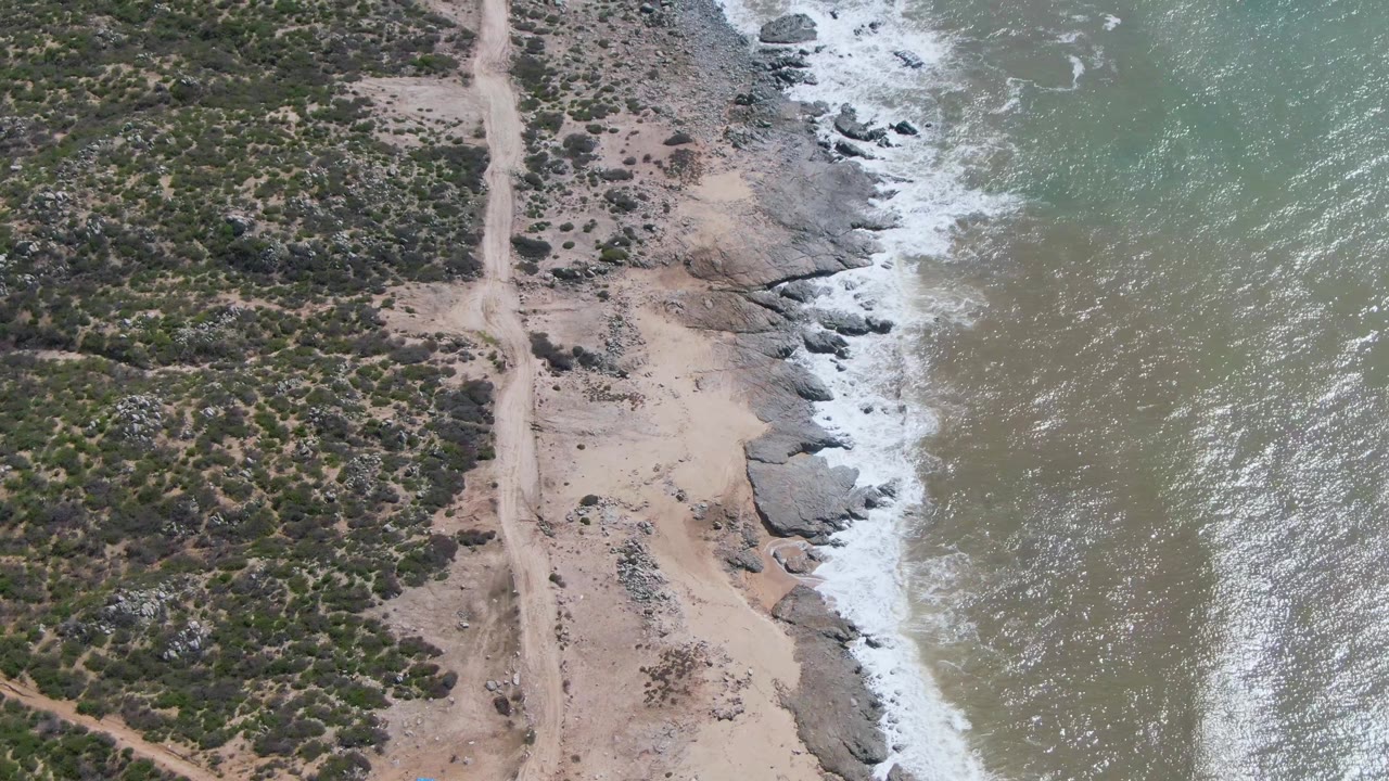 Overhead coastal road view with ocean surf, rocks, beach sand, and low vegetation.