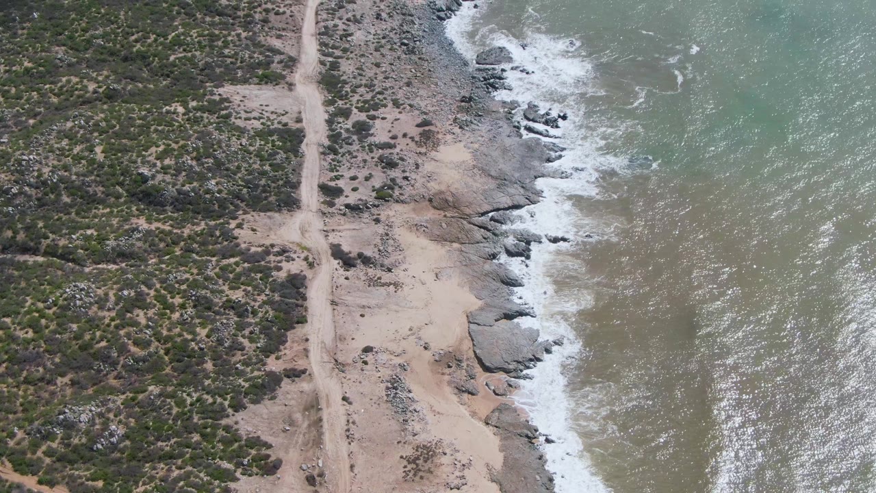 At Buzzards Beach East Cape in Mexico, an aerial frame shows a pale dirt road along scrub-covered land with rocky surf and green-blue water to the right, with no visible people.