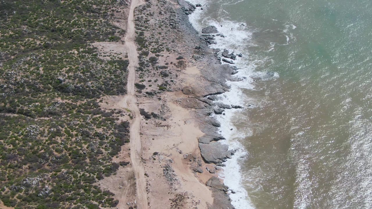 At Buzzards Beach East Cape in Mexico, the road remains near the left center while white foam breaks along the rocks and textured green-blue water fills the right side, with no visible people.