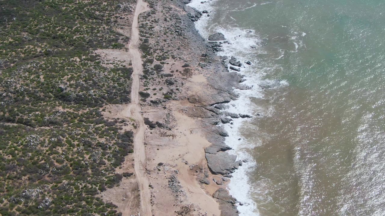 At Buzzards Beach East Cape in Mexico, an overhead view shows scrubland divided by a dirt road, pale sand near the rocks, and foamy surf pushing into the shoreline, with no visible people.