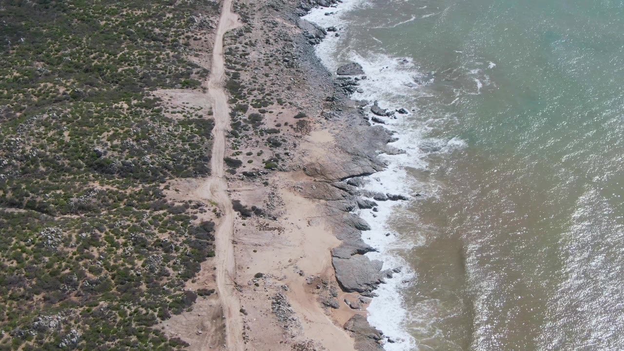Aerial coastline with road, scrub, rocks, and surf wash.
