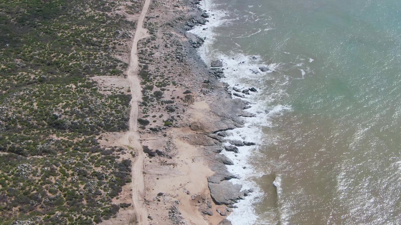Aerial frame over Buzzards Beach East Cape BCS Mexico showing a pale dirt road, scrubland, rocky shore, foamy waves, and no visible people.